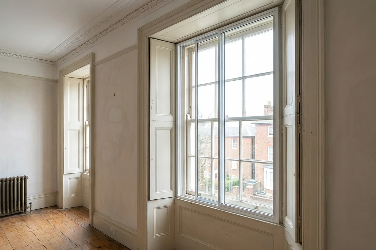 Slim white secondary glazing with two horizontally sliding panes fitted inside the reveal of a tall painted sash window with folding shutters, in a high-ceilinged room with a cornice and bare floorboards