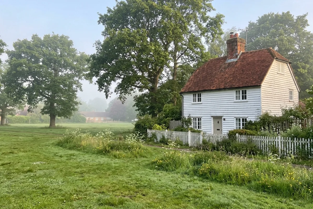 White weatherboarded cottage with a clay tile roof and small casement windows behind a white picket fence beside open grass